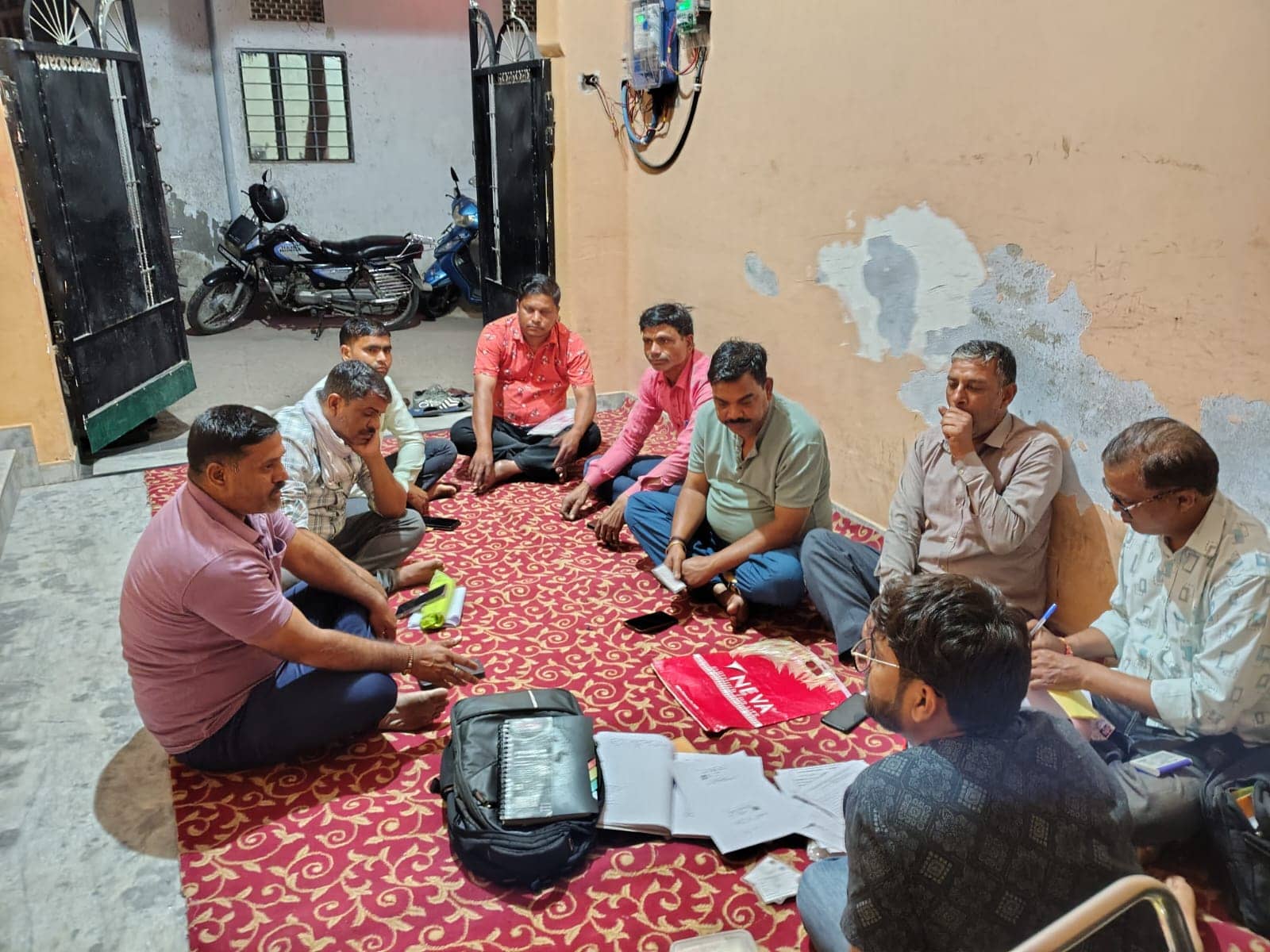 Men sit in a circle on a patterned carpet in an entrance hall at night, with papers, a notebook and bags in the centre.