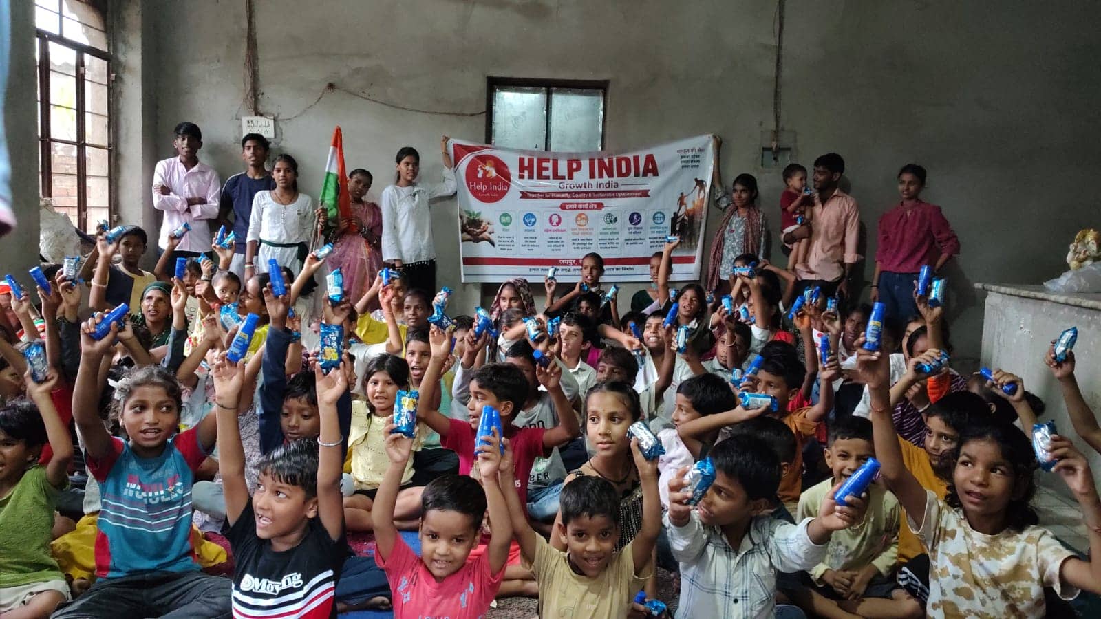 Dozens of smiling children sit on the floor holding up small packets, with young volunteers standing behind them beside a Help India Growth India banner.