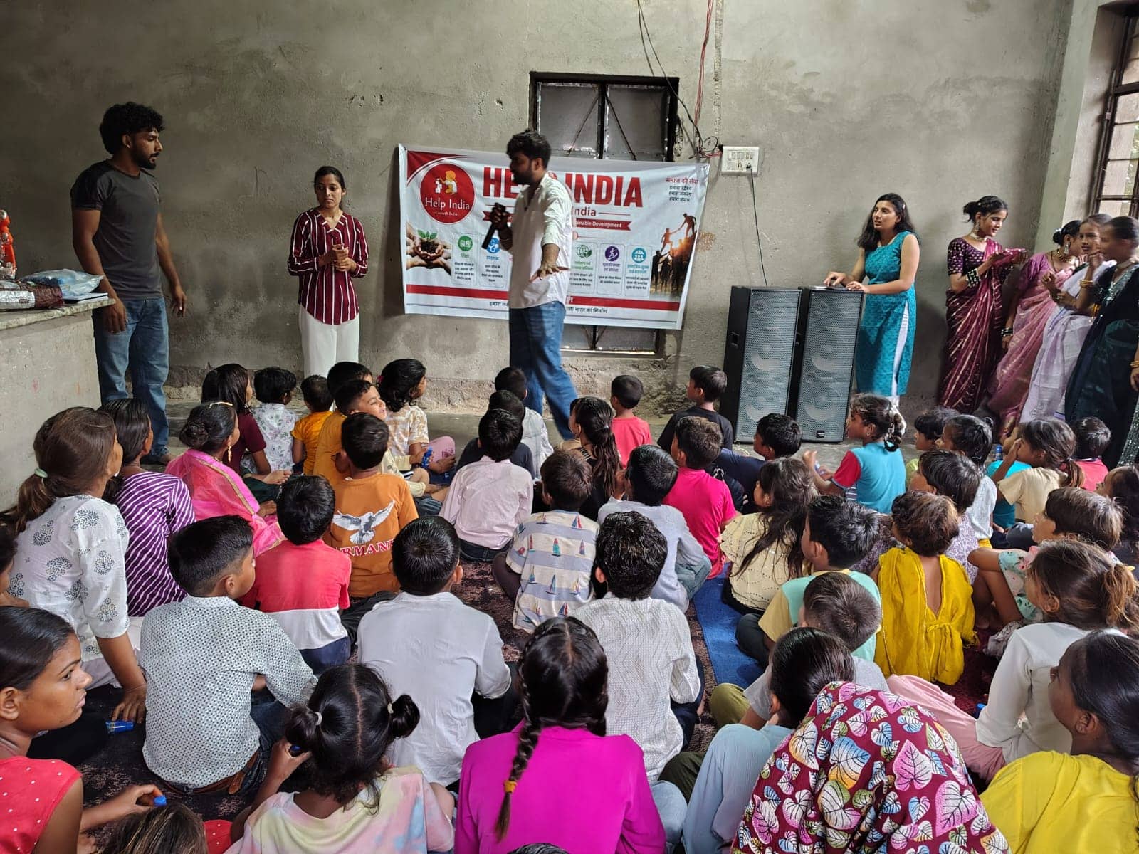 Volunteers speak to a room of seated children, with a Help India Growth India banner on the wall behind them.
