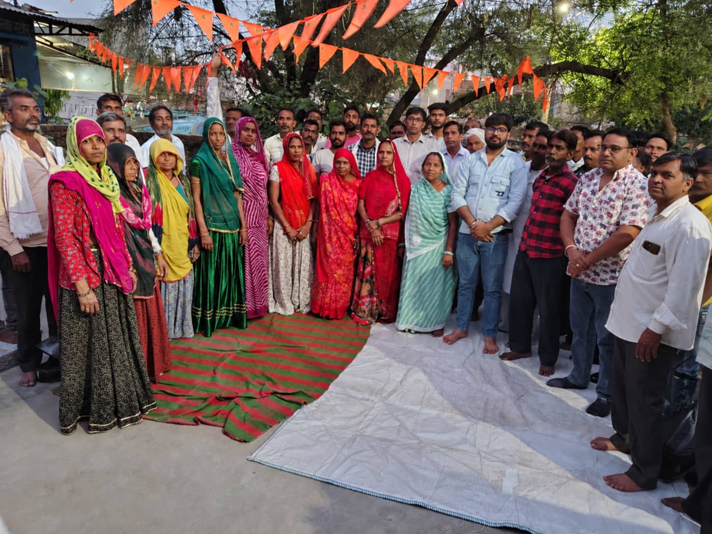 A group of women in colourful Rajasthani dress standing with men from the community, with folders and application papers laid out on a mat in front of them.