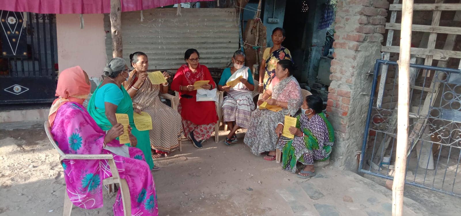 Older women seated on plastic chairs outside a house read yellow information leaflets together.