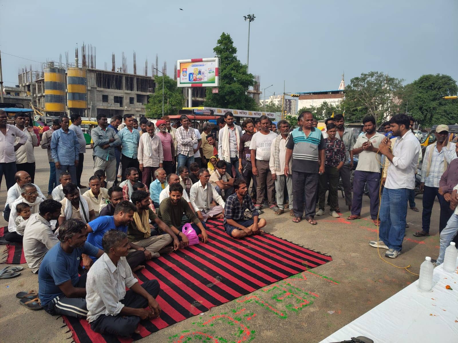A speaker with a microphone addresses a large outdoor gathering of working men, some seated on a red-and-black striped durrie, others standing.